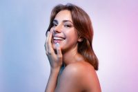 Portrait of smiling woman looking at camera with happiness. Beautiful lady touching face with hand. Wonderful model with short brunette hair posing in studio. Skincare and spa concept